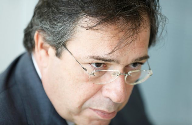 Homem de terno, cabelos grisalhos e óculos, olhando para baixo com foco.