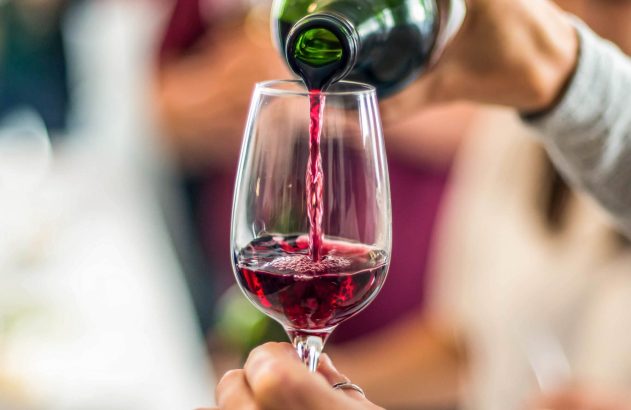 Mão derramando vinho tinto de garrafa verde em taça de vinho, segurada por outra mão.