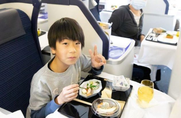 Menino sorridente em assento de avião, comendo refeição e fazendo sinal de paz.