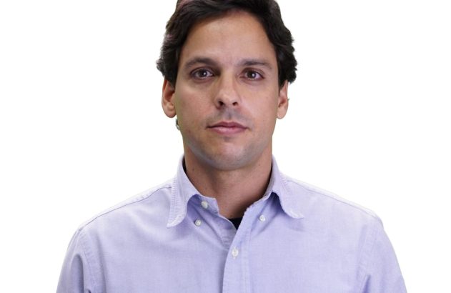 Homem de cabelo escuro e camisa social lilás olhando para frente em fundo branco.