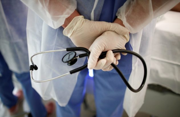 Mãos enluvadas de profissional de saúde seguram um estetoscópio. Veste uniforme azul e avental de proteção branco.
