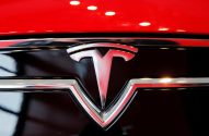 Logotipo da Tesla prateado com detalhes em vermelho, montado na frente de um carro vermelho.