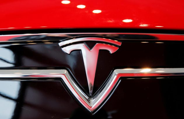 Logotipo da Tesla prateado com detalhes em vermelho, montado na frente de um carro vermelho.