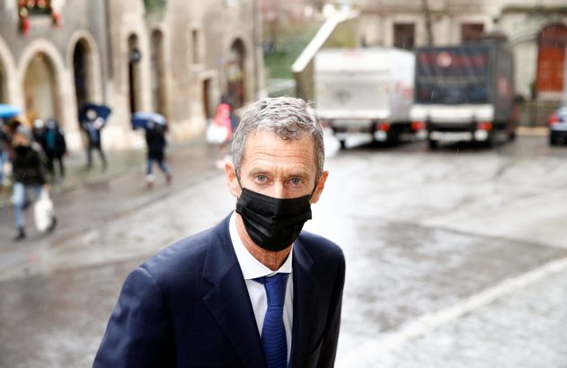 Homem de terno, máscara preta e cabelos grisalhos na chuva, olhando para câmera. Pessoas e veículos borrados ao fundo.