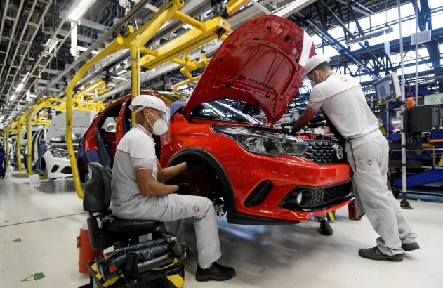 Operários montando um carro vermelho numa linha de produção. Um trabalha no motor (capô aberto), outro na parte frontal.