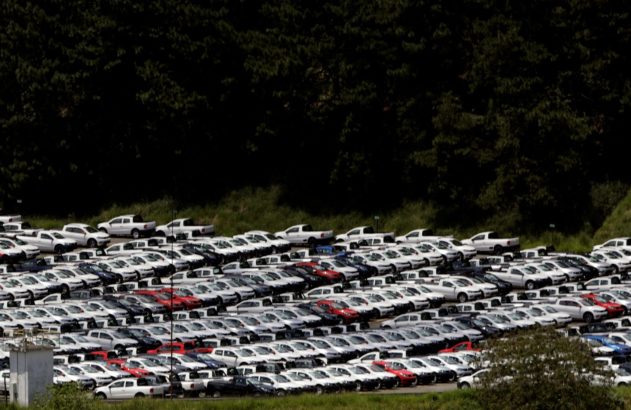 Pátio aberto com centenas de carros novos, em sua maioria brancos, estacionados em fileiras. Ao fundo, uma floresta densa.