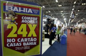 Cartaz promocional da Casas Bahia em evento, com destaque para "24x no Cartão" e pessoas ao fundo.