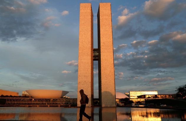 Silhueta de pessoa caminhando à frente do Congresso Nacional em Brasília, refletido na água ao pôr do sol.