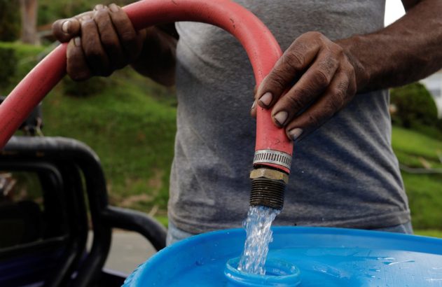 Pessoa enchendo um barril azul de plástico com água jorrando de uma mangueira vermelha.