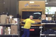 Trabalhador dos Correios empurra carrinho com caixas em galpão, caminhão dos Correios ao fundo.