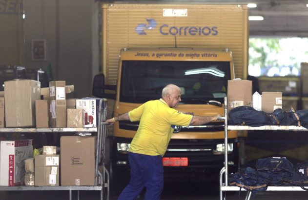 Trabalhador dos Correios empurra carrinho com caixas em galpão, caminhão dos Correios ao fundo.