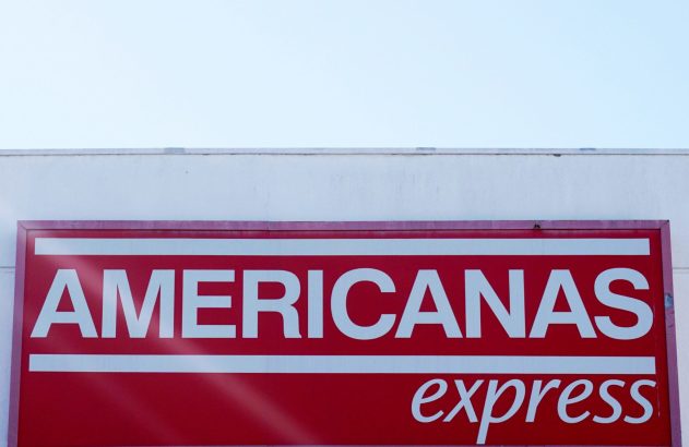 Letreiro vermelho "AMERICANAS express" em prédio branco sob céu azul.
