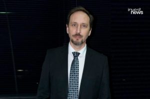 Homem de terno e gravata, olhando para a câmera, com fundo escuro.