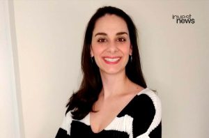 Mulher de cabelo escuro, sorrindo, em suéter preto e branco. Logo 'invest news' no canto superior direito.