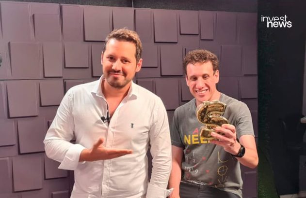 Dois homens em um estúdio, um apontando e o outro sorrindo e segurando um troféu dourado em forma de "S".