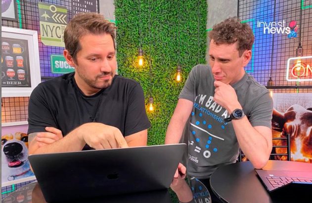 Dois homens olhando para um laptop em uma mesa; um aponta para a tela, o outro observa pensativo.