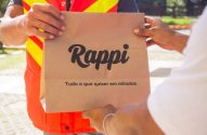 Mãos de entregador Rappi, com colete, passando sacola "Rappi: Tudo o que quiser em minutos" para cliente.