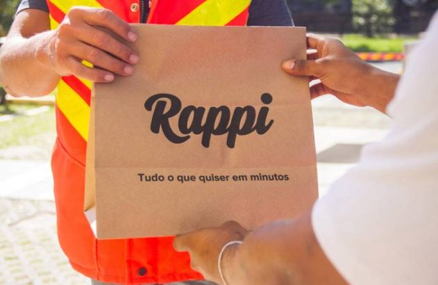 Mãos de entregador Rappi, com colete, passando sacola "Rappi: Tudo o que quiser em minutos" para cliente.