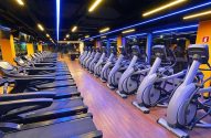 Interior de academia moderna com fileiras de esteiras e elípticos. Iluminação azul e amarela no teto.