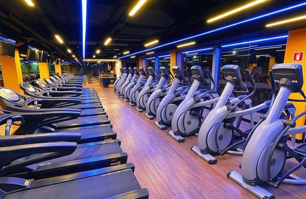 Interior de academia moderna com fileiras de esteiras e elípticos. Iluminação azul e amarela no teto.