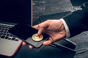 Mão segura smartphone com Bitcoin dourado sobre a tela. Laptop e caneta em mesa.