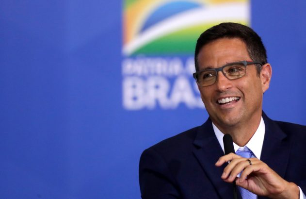 Homem de óculos e terno, sorrindo e falando ao microfone. Fundo azul com logo do Brasil.