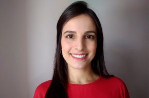 Mulher de cabelos escuros e blusa vermelha sorrindo, com fundo cinza.