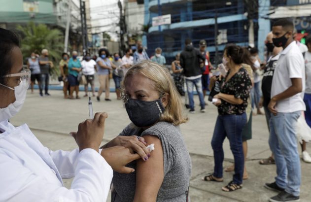 Vacinação: profissional de saúde aplicando injeção em mulher. Pessoas com máscaras no fundo.