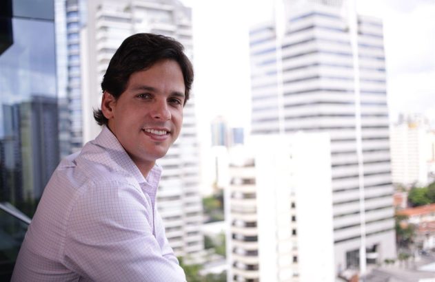 Homem sorrindo para a câmera em sacada, com prédios e paisagem urbana desfocados.
