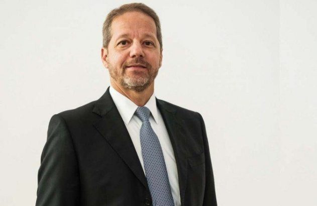 Homem de terno preto, camisa branca e gravata azul estampada, olhando para frente. Fundo branco.