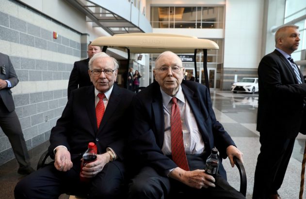 Dois homens idosos de terno sentados em um carrinho, segurando refrigerantes, em um saguão.