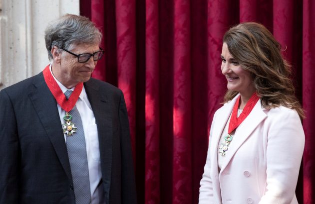 Bill e Melinda Gates, com condecorações no pescoço, se olham sorrindo contra um fundo vermelho.