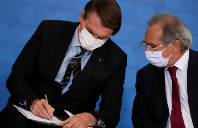 Homem de terno e máscara branca assina documento, outro o observa. Fundo azul.