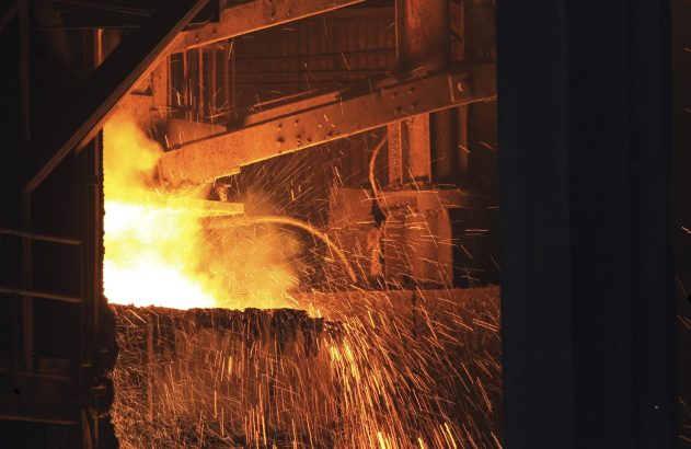 Metal incandescente derramando em ambiente industrial, com jatos de faíscas e fumaça.