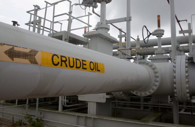 Um duto industrial cinza com rótulo "CRUDE OIL" e seta. Válvulas e tubulações visíveis em uma instalação de petróleo.
