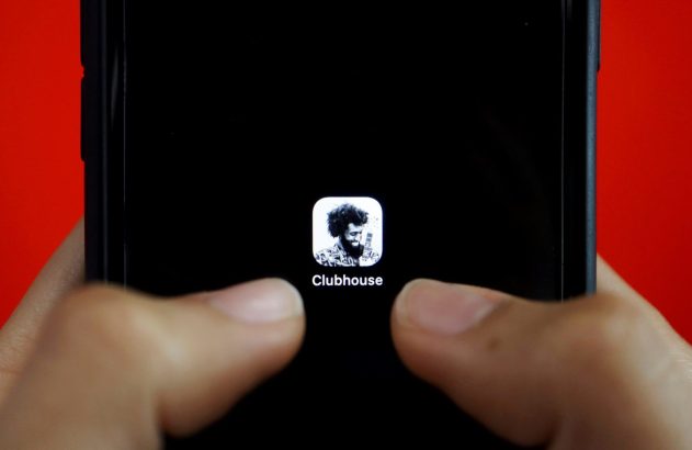 Mãos seguram smartphone, app Clubhouse em destaque na tela preta. Fundo vermelho.