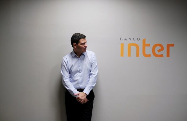 Homem de camisa azul posa ao lado do logo "Banco Inter" em parede cinza, olhando para a esquerda.