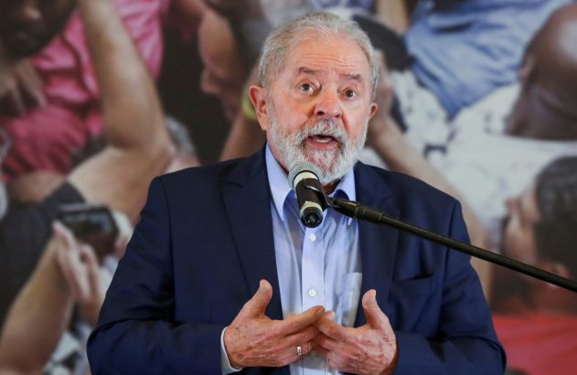 Senhor de barba grisalha discursando ao microfone, vestindo terno azul. As mãos estão em gesto. Fundo desfocado com pessoas.