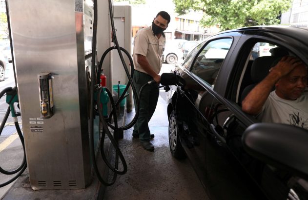 Frentista com máscara abastecendo carro preto em posto de gasolina, motorista dentro do veículo.