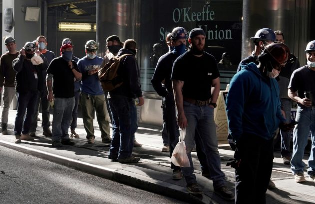 Trabalhadores com capacetes e máscaras aguardando em uma calçada.