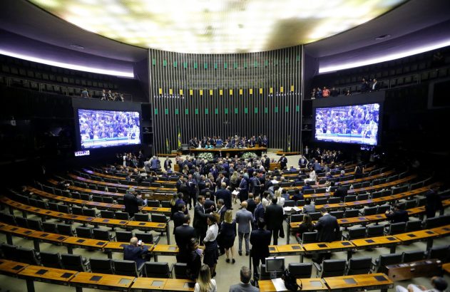 Visão elevada de sessão em plenário legislativo. Políticos conversam e telas exibem o evento.