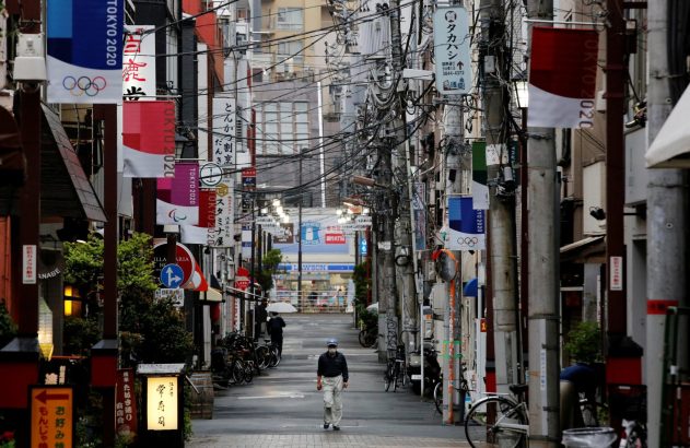 Homem de máscara caminha em rua estreita de Tóquio com banners das Olimpíadas 2020 e fios elétricos emaranhados.