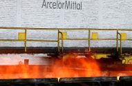 Metal incandescente sendo processado na ArcelorMittal, com grades amarelas e parede de tijolos ao fundo.