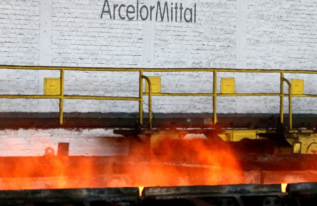 Metal incandescente sendo processado na ArcelorMittal, com grades amarelas e parede de tijolos ao fundo.