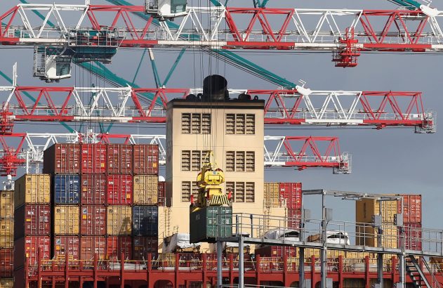 Cena de porto industrial: guindastes enormes carregam um navio de contêineres, com um contêiner verde sendo içado. Contêineres empilhados.