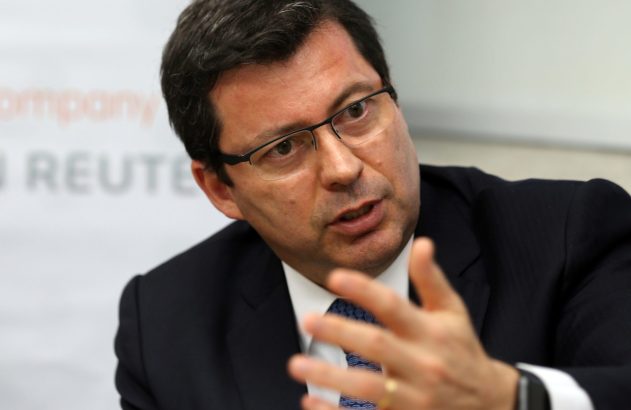 Executivo de terno e óculos gesticula, falando. Fundo com logotipo Reuters desfocado.