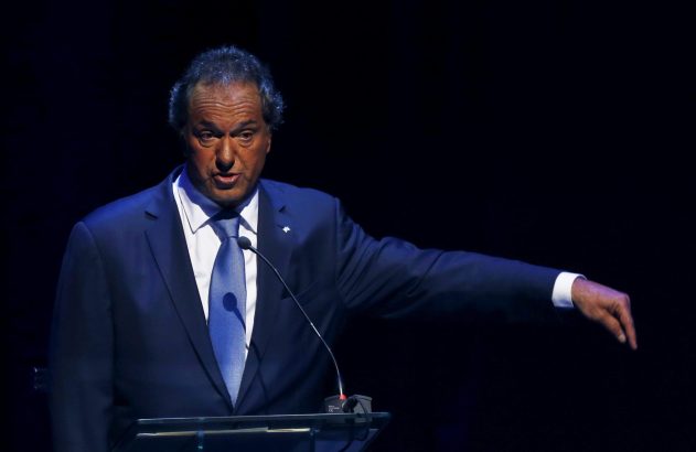 Homem em terno azul e gravata, falando em pódio com microfone, gesticula com braço estendido para o lado.