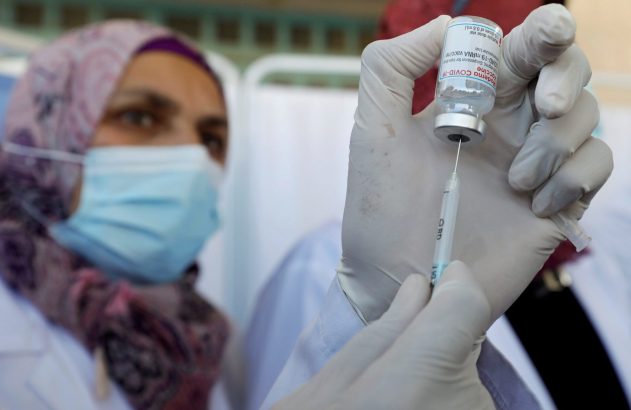 Mãos enluvadas aspiram vacina Moderna COVID-19 de frasco para seringa; pessoa com máscara ao fundo.