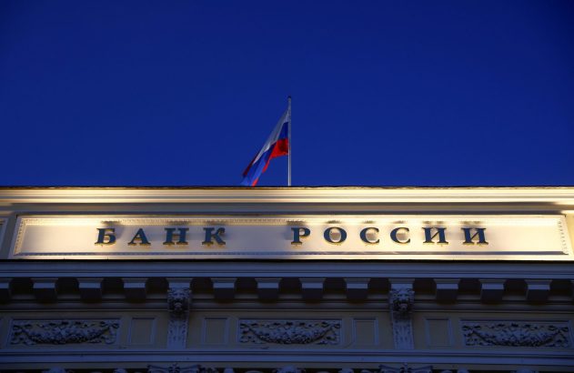 Letreiro 'Banco da Rússia' iluminado em prédio, bandeira russa e céu azul-escuro.
