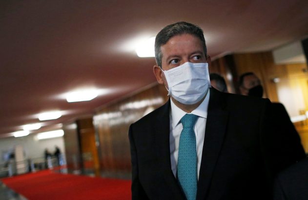 Homem de terno e máscara branca, olhando para o lado em corredor com tapete vermelho.